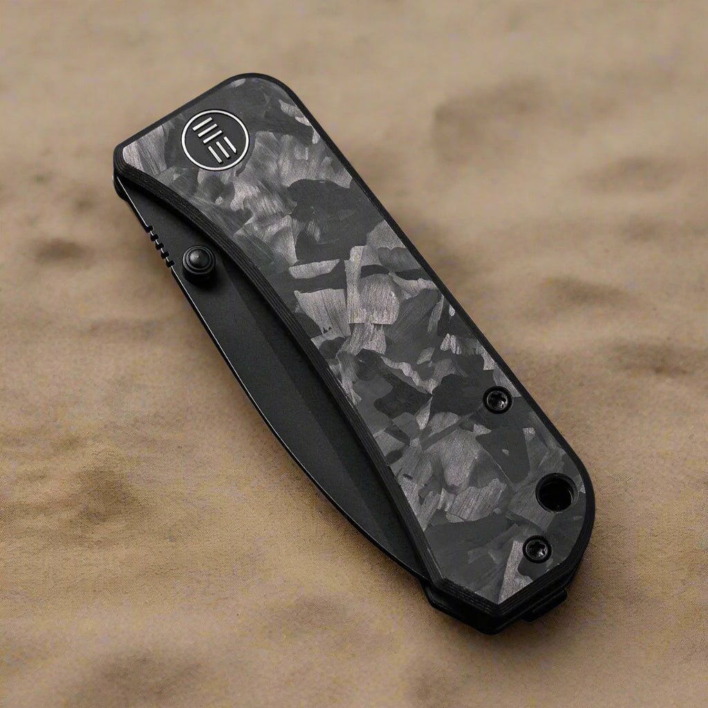 folded knife Banter Marble Carbon laying on beach sand, showing WE logo, carbon pattern design, and thumbstud